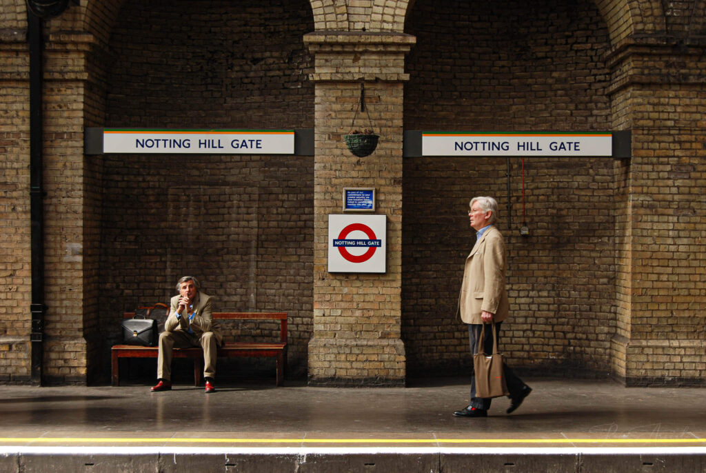 Tube station platform in London. One man is sitting waiting for the train, another man walking across the platform with his suitcase